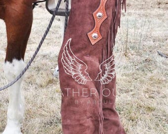 Op de afbeelding: Bruine suède leren chaps met decoratieve zilveren klinknagels en franjes. De chaps worden gedragen met een bruine leren riem en een blauwe spijkerbroek. De afbeelding toont ook een paard.