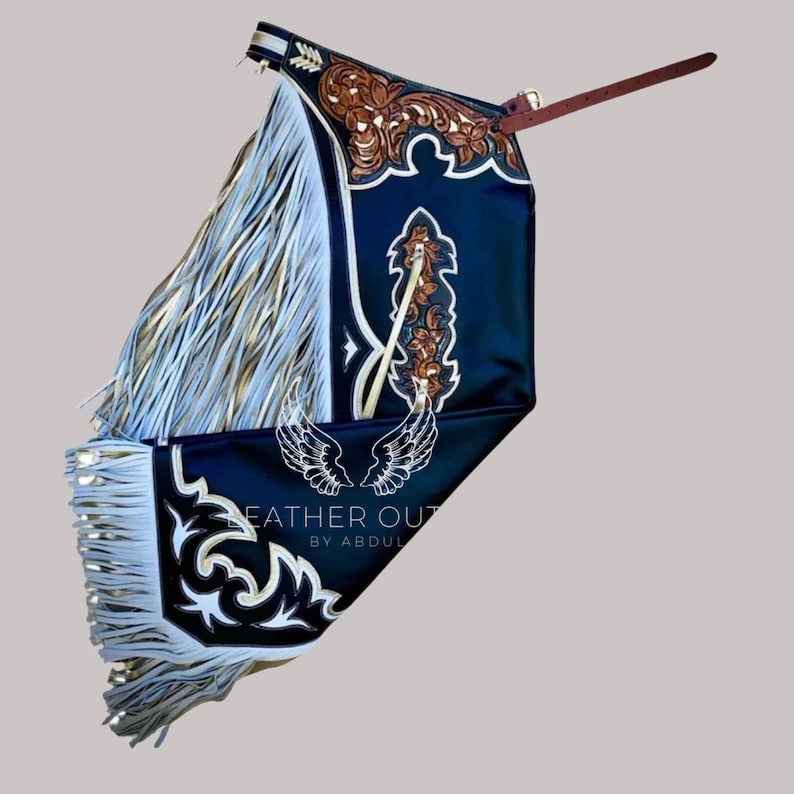 May include: A black leather chaps with white and gold fringe. The chaps feature intricate floral and scrollwork designs in brown and white. The text "LEATHER OUT BY ABDUL" is visible on the bottom of the chaps.