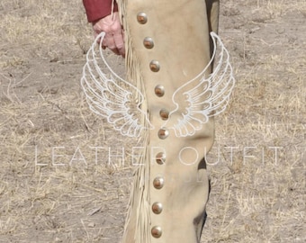 Op de afbeelding: Tan leren chaps met decoratieve metalen studs en franjes langs de zijkanten. De chaps worden gedragen met een bruine leren riem en een zilveren gesp. De afbeelding toont een persoon die de chaps draagt.