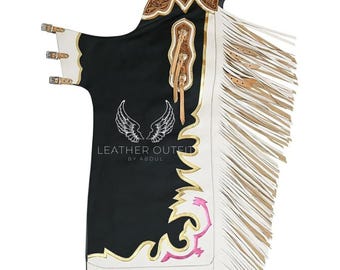 Op de afbeelding: Zwarte leren chaps met witte en gouden accenten, met franjes langs de zijkant. De chaps hebben een decoratieve bovenkant met bloemmotieven en de tekst "LEATHER OUTFIT BY ABDUL".