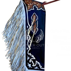 May include: A navy blue leather chaps with intricate brown and white decorative patterns. The chaps feature long, flowing fringe in shades of white and gold. The text "LEATHER OUT" and "ABDUL" is visible.