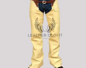 Op de afbeelding: Crèmekleurige leren chaps met bruine leren details en een riem. De chaps hebben een vleugelontwerp op elke been en de tekst "LEATHER OUTFIT BY ABDUL". Ze worden gedragen over een blauwe spijkerbroek en bruine laarzen.