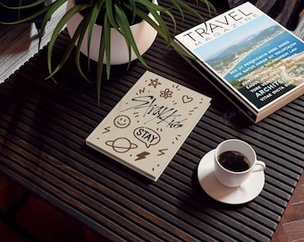 May include: A coffee table with a plant in a white pot, a magazine, a cup of coffee, and a book with the text "Stray Kids STAY" and various doodles. The table is dark brown with a slatted design.