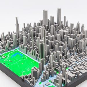 May include: A detailed 3D model of a city, with numerous grey buildings and skyscrapers. A green area, likely a park, is included, providing a contrast. The model is mounted on a black base.