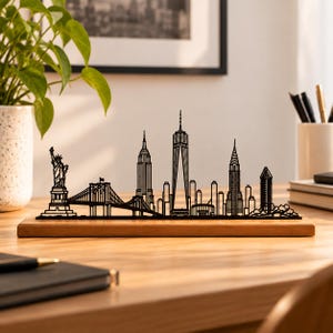 Può includere: Una scultura a silhouette in metallo nero dello skyline di New York montata su una base rettangolare in legno. Il design raffigura la Statua della Libertà, il ponte di Brooklyn e iconici grattacieli cittadini.