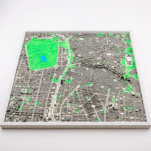 May include: A detailed 3D map of a city, with a grey and green street layout. The map is set within a white, textured frame. Green areas represent parks and open spaces, with a blue area indicating a body of water.