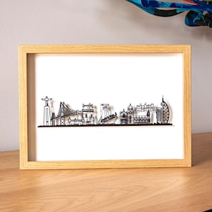 Könnte beinhalten: Ein gerahmtes Schwarz-Weiß-Skyline-Kunstwerk. Das Kunstwerk zeigt verschiedene Gebäude und Wahrzeichen, darunter eine Brücke, ein Schiff und ein Turm. Der Rahmen ist aus hellem Holz gefertigt, und der Hintergrund ist weiß.