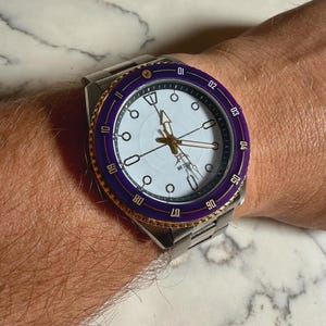 May include: A stainless steel wristwatch with a white face, gold hands, and a purple and gold bezel. The watch has a silver band and is worn on a wrist. The watch face has circular and rectangular markers.