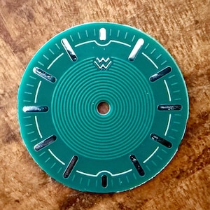May include: A green watch face with white markings and silver accents. The face has a circular pattern and a small hole in the centre.