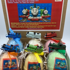 May include: A gift set of six egg-shaped bath bombs with Thomas the Train toys inside. The box includes bath bombs in various colors, each topped with a toy train. The box has text that reads "Thomas the Train Bath Bomb Gift Set".