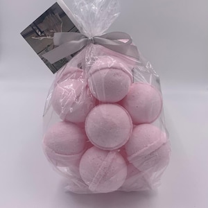 May include: A clear cellophane bag tied with a gray ribbon contains several light pink bath bombs. A small card is attached to the bag. The bath bombs are spherical with a textured surface.