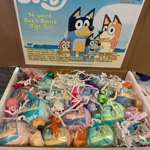 May include: A 14-pack bath bomb gift set featuring Bluey characters. The box contains individually wrapped bath bombs in various colors, tied with ribbons. The box has the text "Bluey" and "Northwoods Bath & Spa".