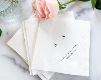 May include: White paper napkins with the initials "A S" and the names "AVEDA AND STEVEN" and the date "Saturday, May 27, 2023" printed in black. A pink rose rests on top of the napkins. Wedding or event stationery.