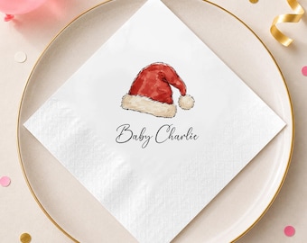 May include: A white square paper napkin featuring a watercolor illustration of a red Santa hat with a white fur trim and pom-pom. Below the hat, the text Baby Charlie is printed in a black cursive script font on the napkin surface.