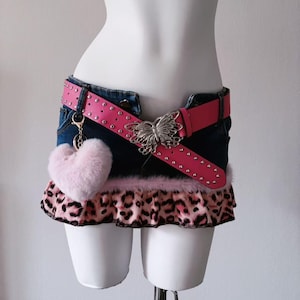 May include: A denim mini skirt with a pink studded belt featuring a butterfly buckle. The skirt has a leopard print ruffle and a pink heart-shaped keychain. The skirt is displayed on a mannequin.