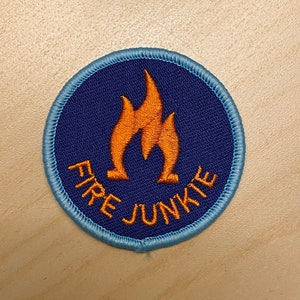 Könnte beinhalten: Ein runder blauer und violetter Patch mit einem gestickten Rand. Der Patch hat ein orangefarbenes Flammenmotiv und den Text "FIRE JUNKIE".