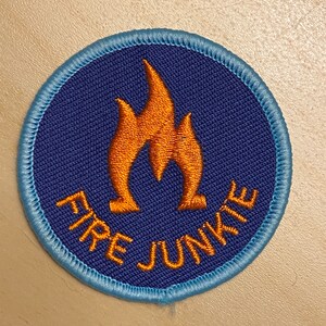K&ouml;nnte beinhalten: Ein runder, gestickter Aufn&auml;her in Blau und Orange mit dem Text "FIRE JUNKIE" und einer stilisierten orangen Flamme in der Mitte.