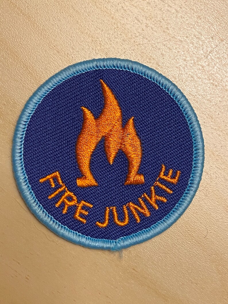 K&ouml;nnte beinhalten: Ein runder, gestickter Aufn&auml;her in Blau und Orange mit dem Text "FIRE JUNKIE" und einer stilisierten orangen Flamme in der Mitte.