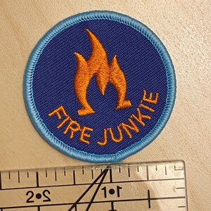 K&ouml;nnte beinhalten: Ein gestickter Aufn&auml;her in Blau und Orange mit dem Text "FIRE JUNKIE" und einem Flammenmotiv.