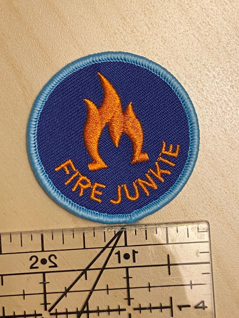 K&ouml;nnte beinhalten: Ein gestickter Aufn&auml;her in Blau und Orange mit dem Text "FIRE JUNKIE" und einem Flammenmotiv.