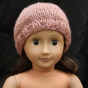 May include: A knitted pink hat with a textured pattern. The hat is worn by a doll with brown eyes and a neutral expression. The hat appears to be handmade and is a soft, muted pink color.