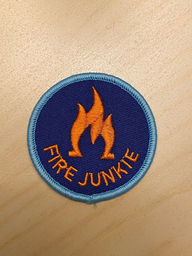 K&ouml;nnte beinhalten: Ein runder blauer und violetter Patch mit einem gestickten Rand. Der Patch hat ein orangefarbenes Flammenmotiv und den Text "FIRE JUNKIE".