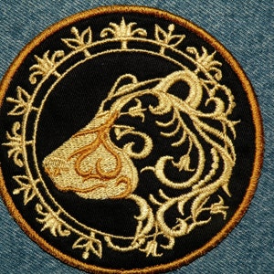 May include: A gold embroidered patch with a black background featuring a stylized lion's head with intricate floral details.