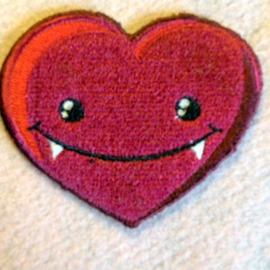 May include: A red felt heart with a smiling face and fangs. The heart has white stitching around the eyes and fangs.