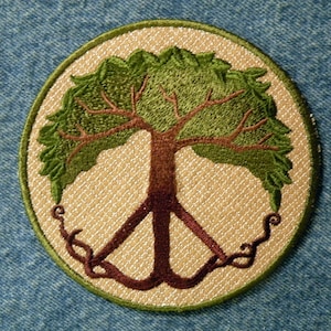 May include: A round embroidered patch with a peace sign and a tree growing out of it. The tree has green leaves and a brown trunk. The peace sign is brown and has vines growing around it.