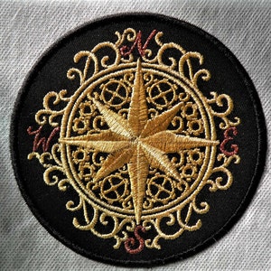 May include: A black embroidered patch with a gold compass design. The compass features a star with four points and a circular design with intricate details. The compass is surrounded by a decorative border.
