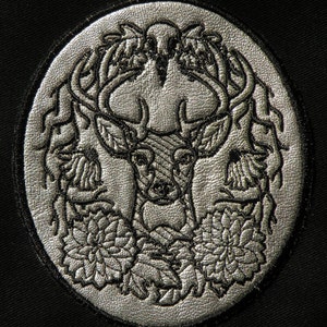 May include: A circular, silver-colored embroidered patch featuring a detailed stag design. The stag has large antlers and is surrounded by floral and leaf motifs. The patch is set against a black background, highlighting the intricate details of the embroidery.