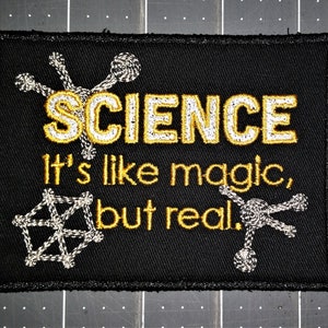 Könnte beinhalten: Schwarzer rechteckiger Aufnäher mit dem Wort "SCIENCE" in goldener Stickerei und dem Satz "It's like magic, but real." in goldener Stickerei. Der Aufnäher zeigt gestickte Molekulardesigns in Weiß und Gold.