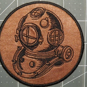May include: A brown leather patch with a black embroidered image of a vintage diving helmet. The helmet has a large round window and a small round window on the side. The helmet is attached to a hose and a breathing apparatus.