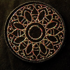 May include: A close-up of a circular, embroidered design on a dark brown leather surface. The design features a central circle surrounded by intricate, multi-colored floral patterns.