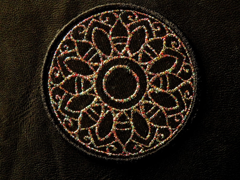 May include: A close-up of a circular, embroidered design on a dark brown leather surface. The design features a central circle surrounded by intricate, multi-colored floral patterns.