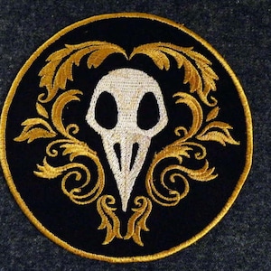 May include: A black and gold embroidered patch featuring a stylized white skull with a beak, surrounded by ornate floral designs.