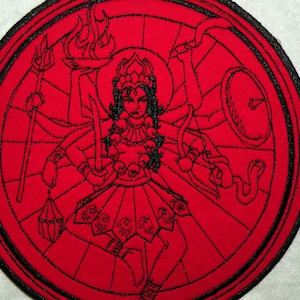 May include: A red embroidered patch with a black outline depicting a deity with multiple arms holding various objects, including a trident, a sword, and a shield. The deity is surrounded by a circular design with flames and skulls.
