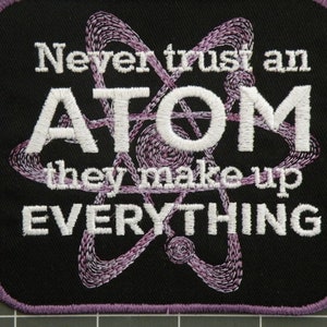 Könnte beinhalten: Ein schwarzer gestickter Aufnäher mit einem violetten Atomdesign und dem Text "Never trust an ATOM they make up EVERYTHING".
