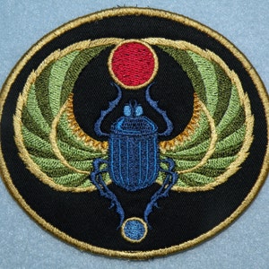 May include: A circular embroidered patch with a gold border, depicting a blue scarab beetle with green wings, a red circle above, and a blue circle below.