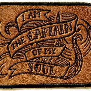 May include: A brown leather patch with black embroidery. The patch features a banner with the text "I AM THE CAPTAIN OF MY SOUL".