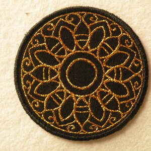 May include: A black embroidered patch with a gold circular design. The design features a central circle surrounded by a ring of intricate floral patterns.