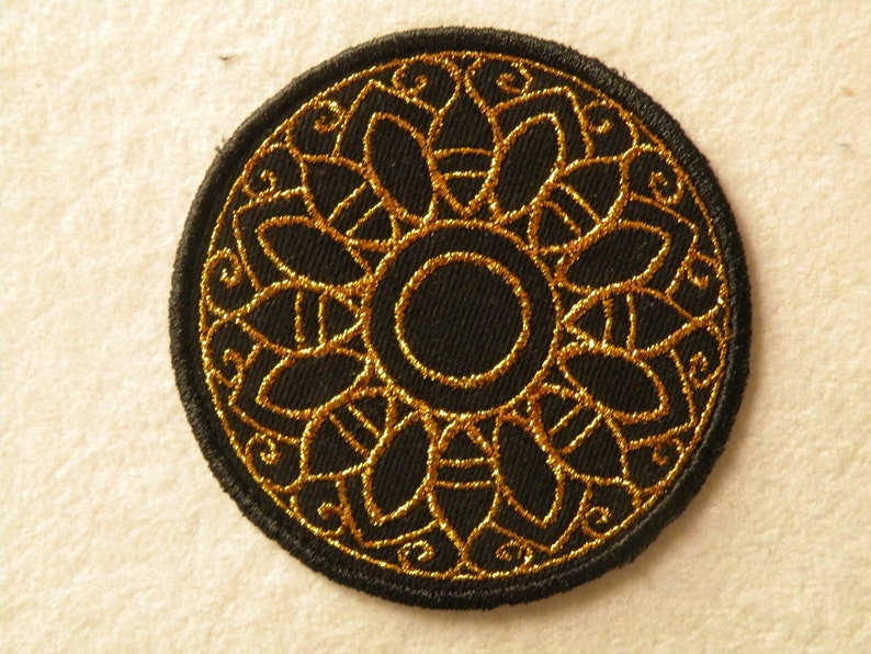 May include: A black embroidered patch with a gold circular design. The design features a central circle surrounded by a ring of intricate floral patterns.