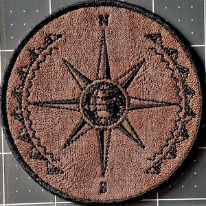 May include: A brown leather patch with a black compass design. The compass has a starburst shape with a globe in the center. The compass is surrounded by a black chain-link design.