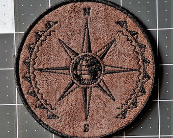 May include: A brown leather patch with a black compass design. The compass has a starburst shape with a globe in the center. The compass is surrounded by a black chain-link design.