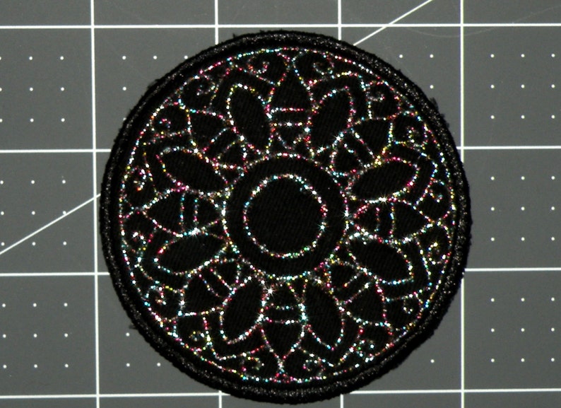 May include: Black round patch with a silver glitter design of a circular mandala with a smaller circle in the center.