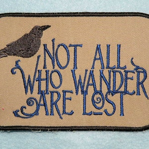 Könnte beinhalten: Ein beigefarbener Stoff-Patch mit schwarzen Nähten und einem blauen gestickten Design. Das Design zeigt einen schwarzen Raben und den Text "Not all who wander are lost."