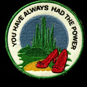 May include: A green and white embroidered patch with the text "You Have Always Had The Power" in a circle. The center of the patch features a green and blue image of the Emerald City from the Wizard of Oz.  Two red ruby slippers are in the foreground.