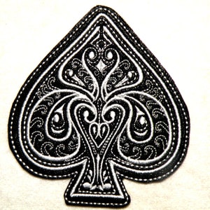 May include: Black and white embroidered patch in the shape of a spade playing card. The spade is decorated with a detailed, intricate design.