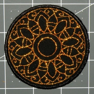 May include: A black and gold embroidered patch with a circular design. The center of the patch features a smaller circle with a black background and gold outline. The outer circle features a repeating pattern of gold leaves and swirls.