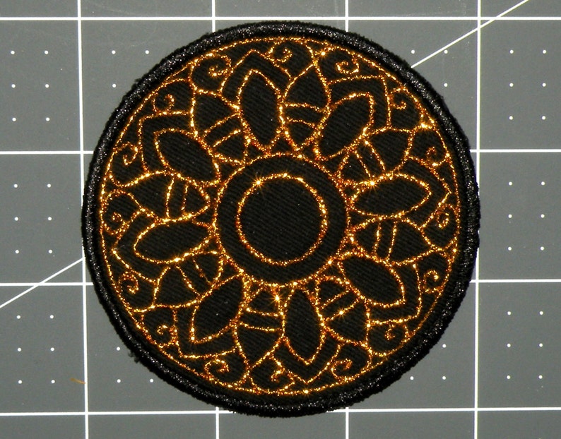May include: A black and gold embroidered patch with a circular design. The center of the patch features a smaller circle with a black background and gold outline. The outer circle features a repeating pattern of gold leaves and swirls.